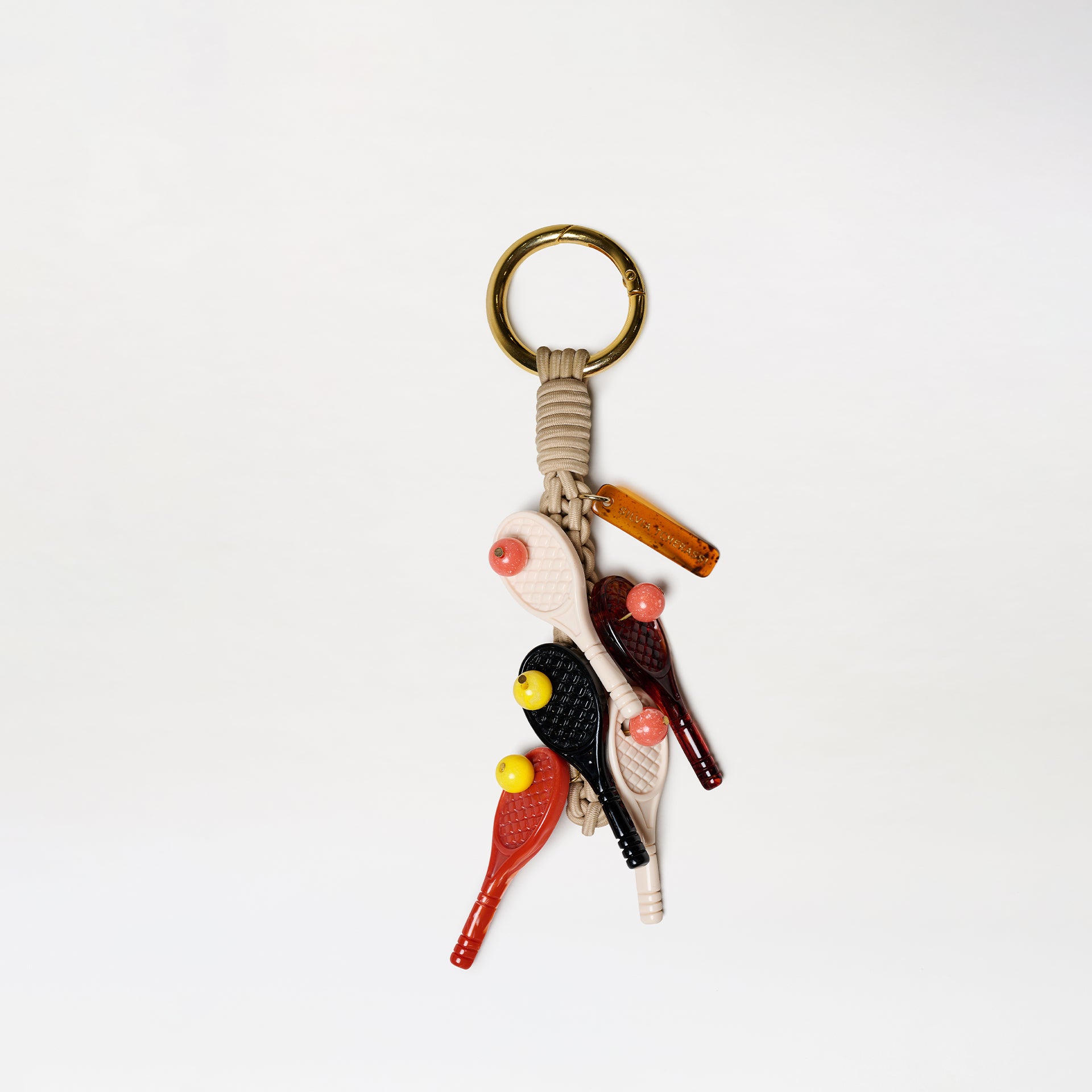 The Tournament Charm keychain features a gold ring, beige cord, and five bird-shaped charms in black, red, and beige with yellow beaks. A small brown rectangular tag is attached. All items are displayed against a white background.