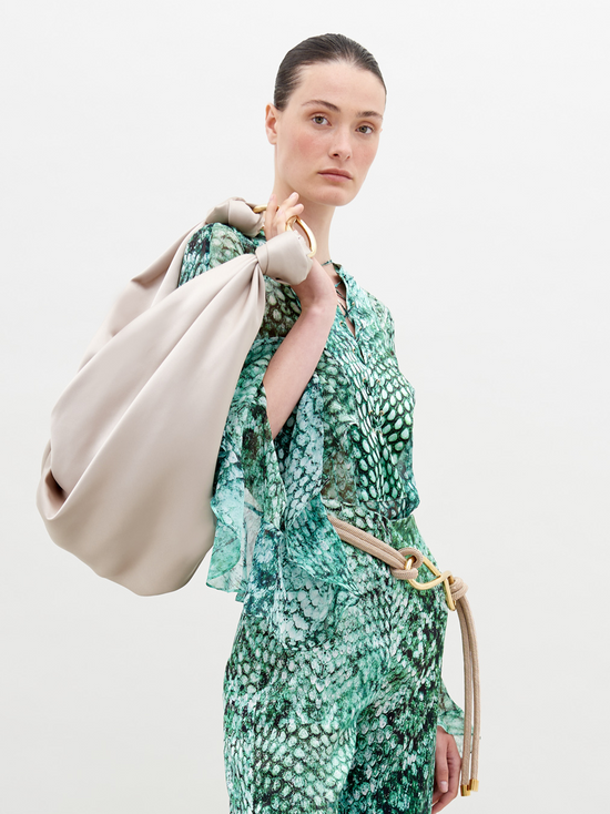 A person with slicked-back hair wears Thea Blouse Emerald Animal Print and holds a large beige bag over their shoulder, perfect for those looking to pre-order now. The background is plain white, highlighting the stylish ensemble expected to ship by November 15th, 2024.