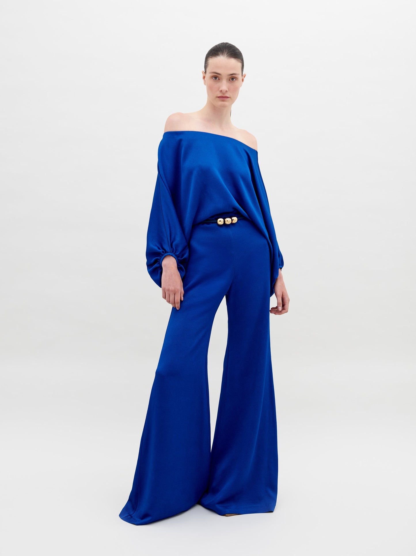 A person stands against a plain background wearing an off-the-shoulder blue top with wide sleeves and matching wide-leg pants, accessorized with a belt that has two large gold embellishments. Pre-order the Theresa Jumpsuit Royal Blue now to ensure you receive your outfit by the ship by date of November 15th, 2024.