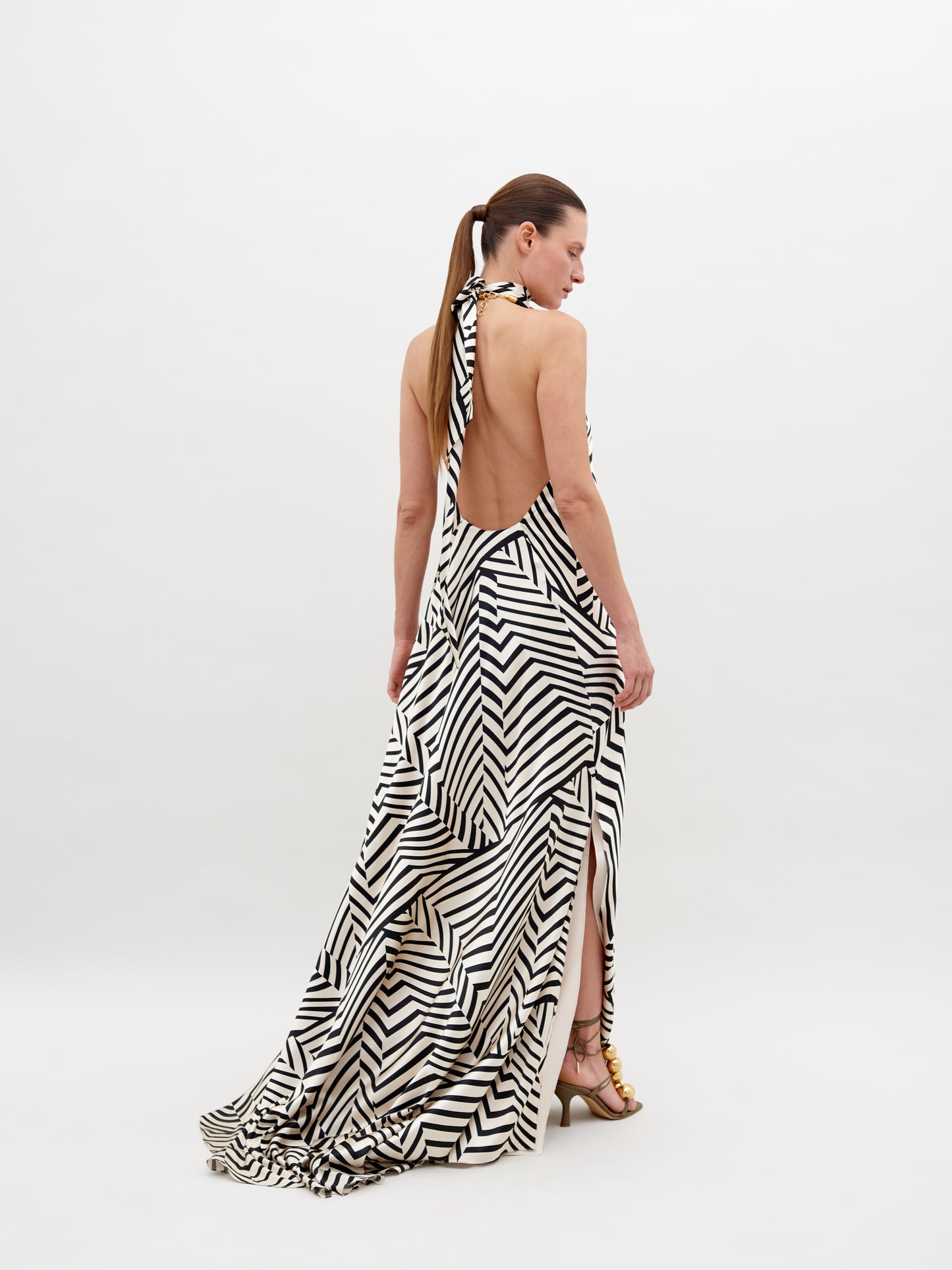 A person stands wearing a Tila Dress Beige Black Stripes with a high neckline, accessorized with a large floral detail at the shoulder, and gold footwear. This stunning outfit is available for pre-order and will ship by November 15th, 2024.