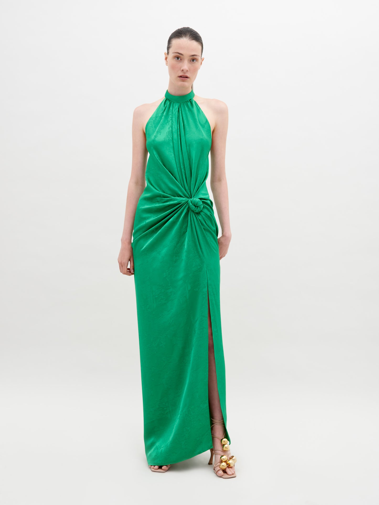 A woman stands against a plain background wearing a sleeveless, high-neck green dress with a front knot and a thigh-high slit. She has her hair pulled back and wears open-toed gold sandals. The Tizziana Dress Green is available for pre-order starting November 15th.