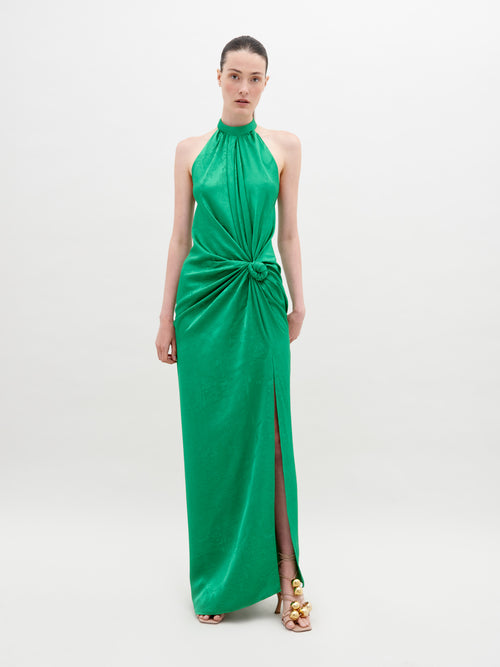 A woman stands against a plain background wearing a sleeveless, high-neck green dress with a front knot and a thigh-high slit. She has her hair pulled back and wears open-toed gold sandals. The Tizziana Dress Green is available for pre-order starting November 15th.