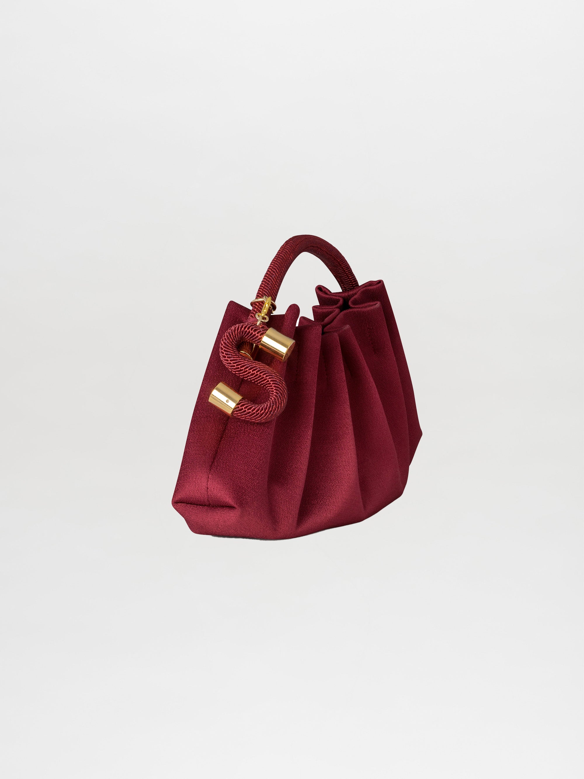 The Trapezius Clutch is a maroon pleated crepe handbag with gold accents, a textured handle, and a gold letter "S" charm.