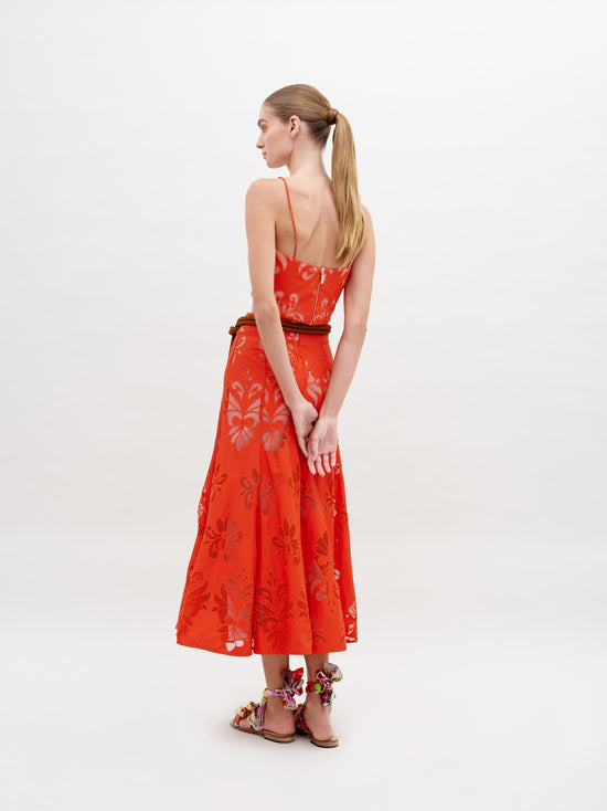 A woman stands wearing a Grettel Skirt Rouge Embroidery with a bright orange floral-patterned two-piece outfit and colorful sandals. She poses against a plain white background, available for pre-order starting November 15th for the 2024 collection.