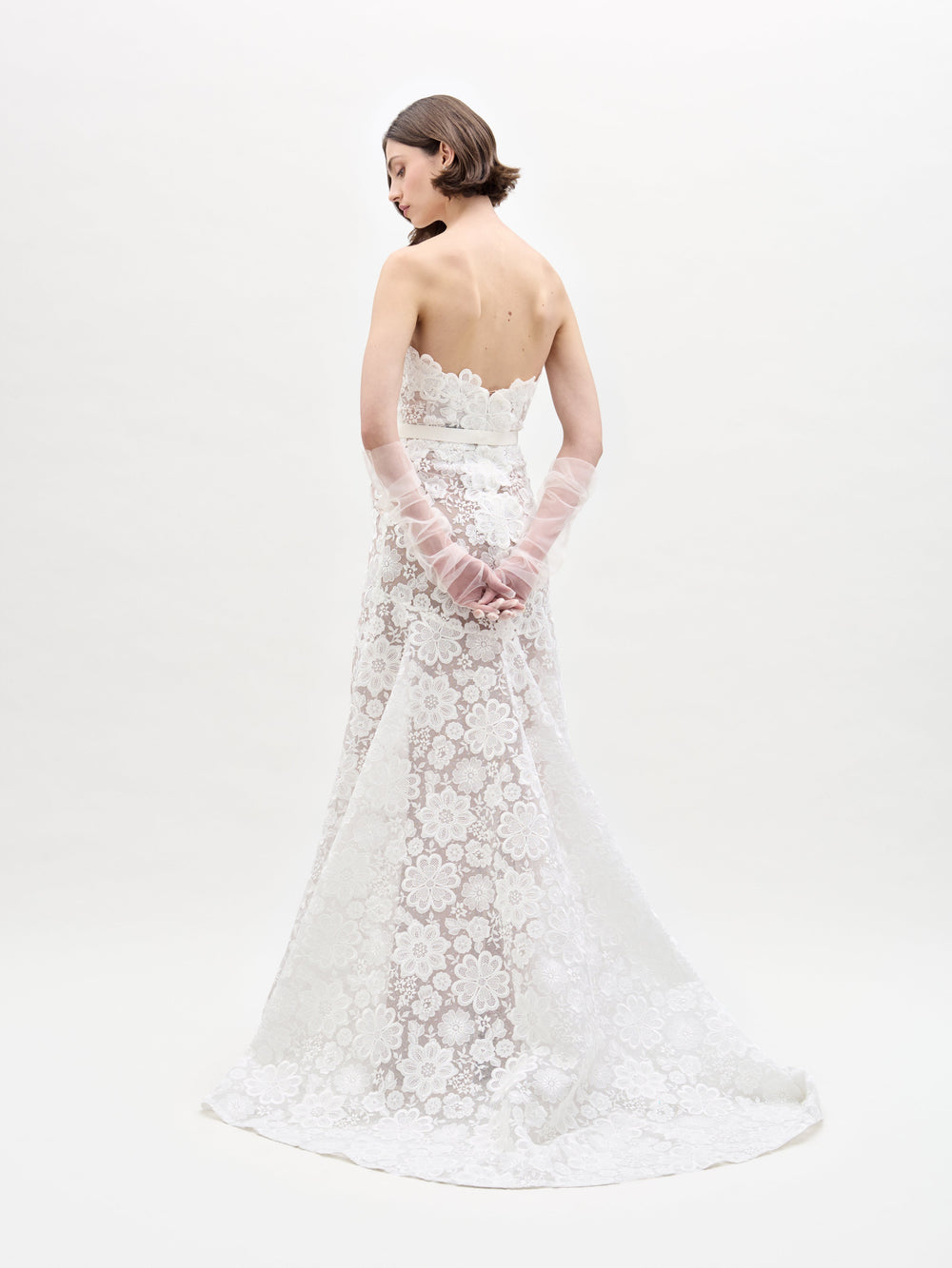 A woman poses against a plain white background in the Valencia Dress, which features a strapless design with lace floral patterns. She complements it with sheer gloves.