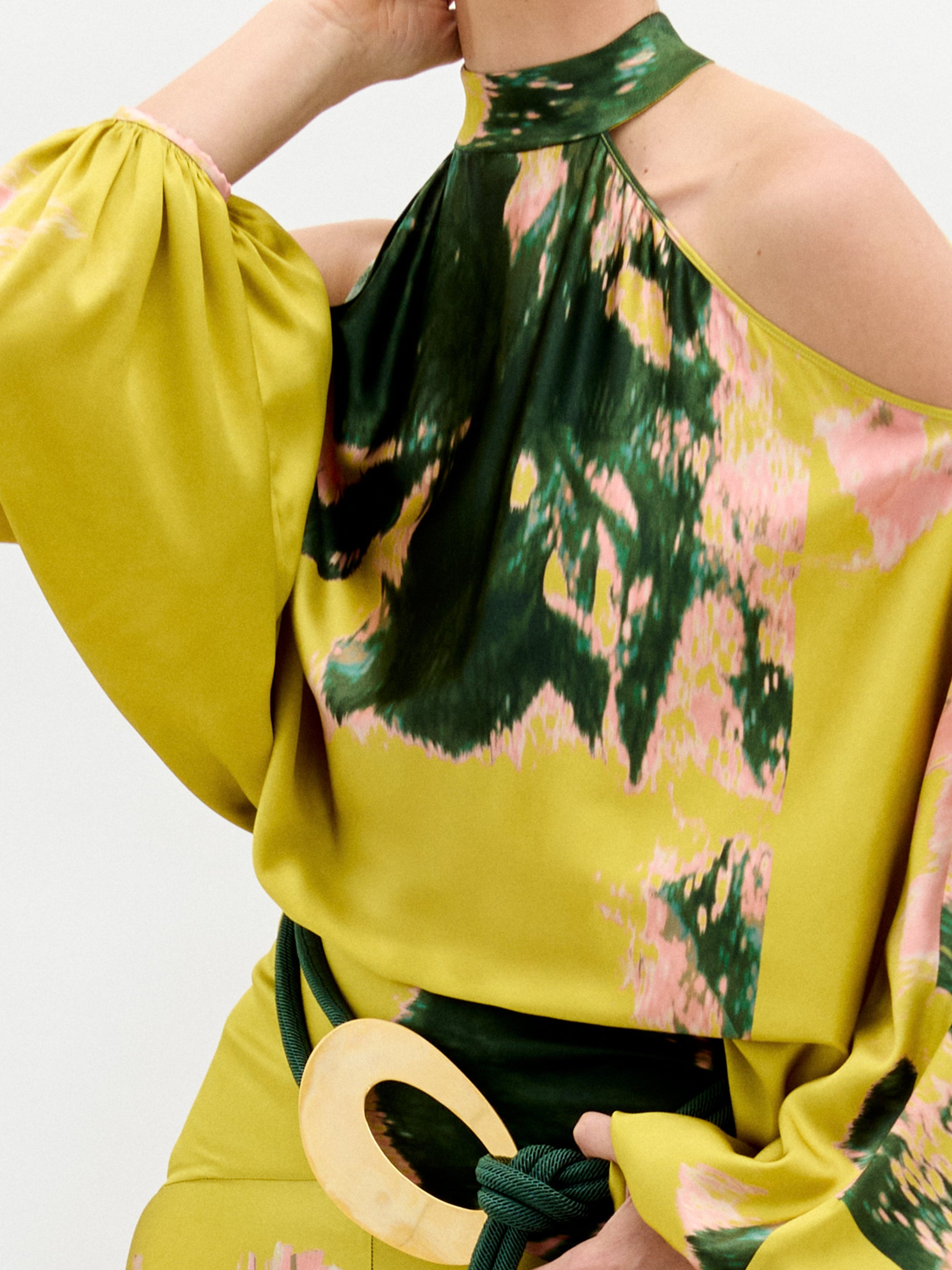 A person wears the Valeria Dress, a flowing maxi dress in yellow, pink, and green patterns with open shoulders and a 3D cord belt, standing against a plain white background.