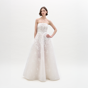 A woman poses against a white background wearing the Viviana Dress, a strapless, floor-length maxi made from Italian fabric with floral embroidery and a fitted bodice, standing with one hand on her hip.