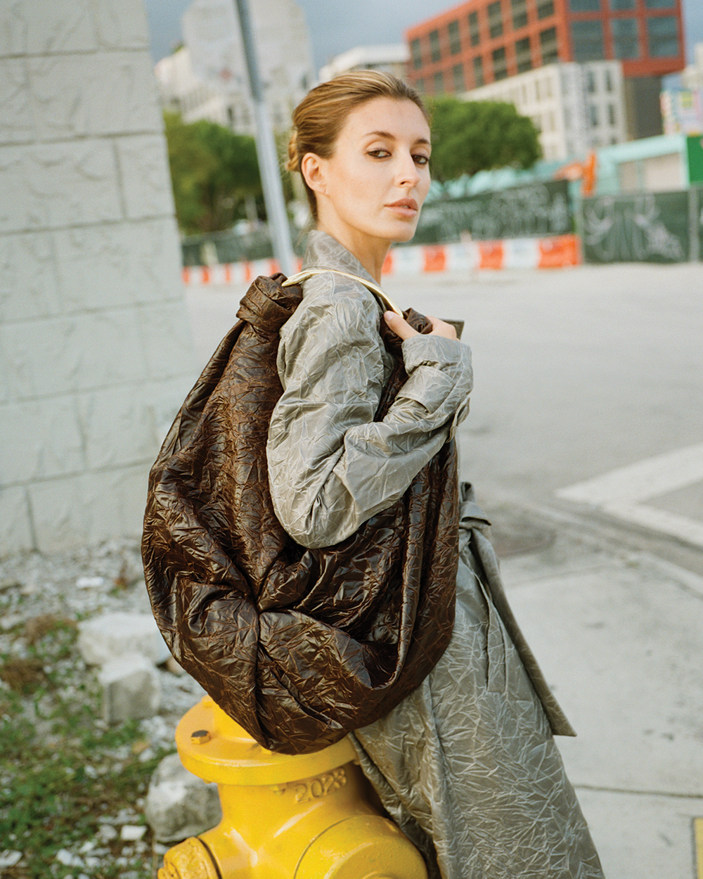 Woman holding a large brown leather jacket outdoors with buildings in the background