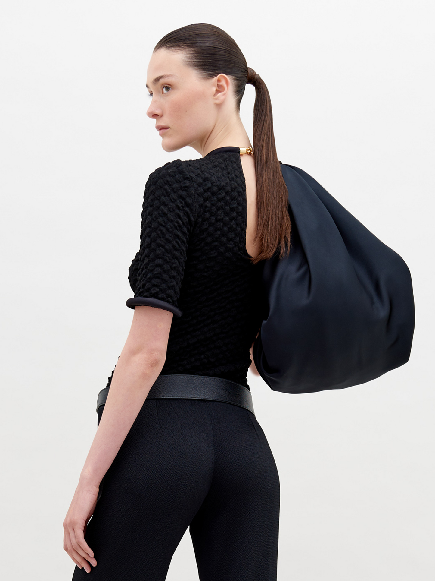 A woman with tied-back hair wears a Venecia Tshirt Black, black pants, and a modern belt, holding a large black bag. She stands against a plain white background, ready to pre-order her outfit by November 15th.