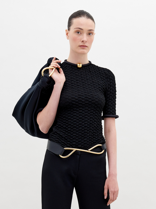 A woman with tied-back hair wears a Venecia Tshirt Black, black pants, and a modern belt, holding a large black bag. She stands against a plain white background, ready to pre-order her outfit by November 15th.