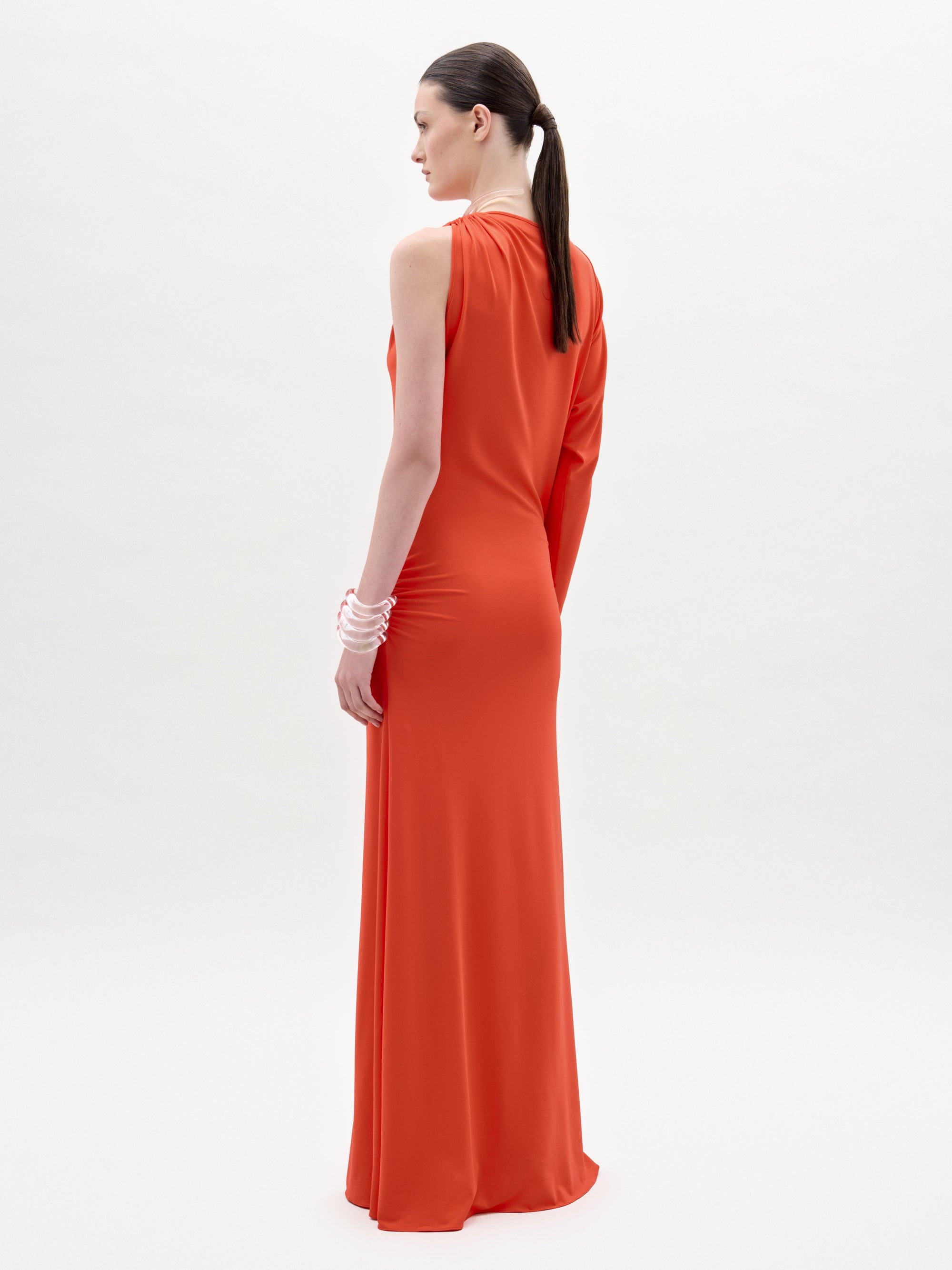 A woman stands against a plain white background, looking directly at the camera while wearing the Virna Dress—a long, one-sleeve orange maxi dress with a sleek column silhouette.