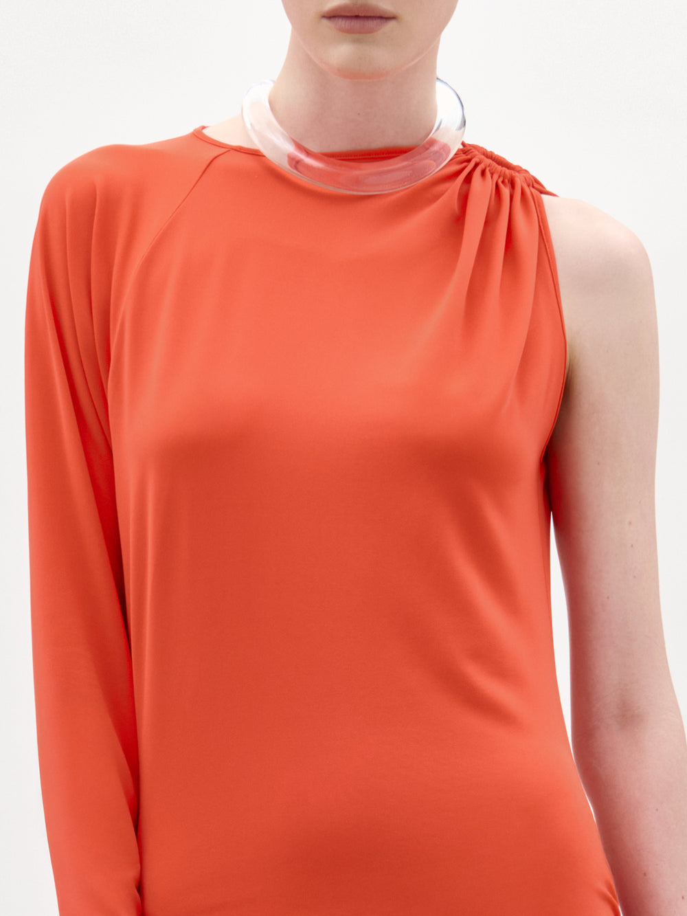 A woman stands against a plain white background, looking directly at the camera while wearing the Virna Dress—a long, one-sleeve orange maxi dress with a sleek column silhouette.