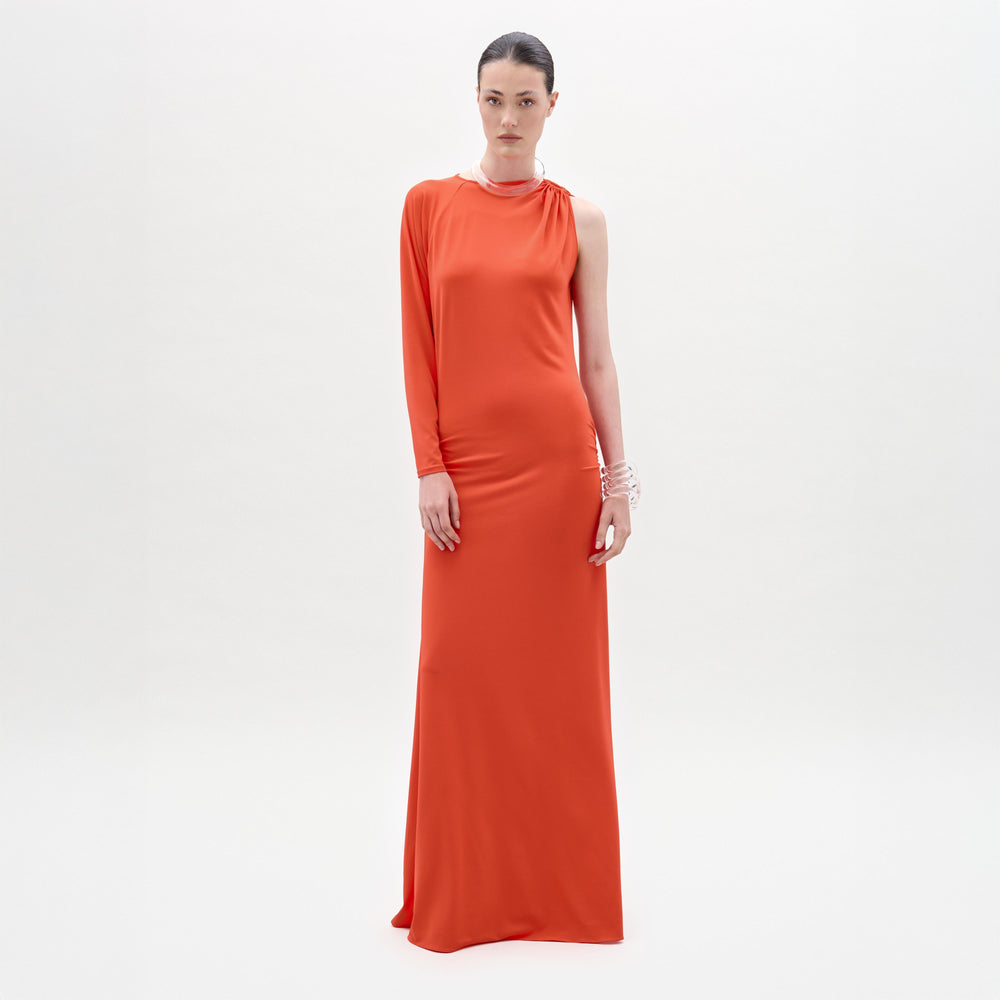 A woman stands against a plain white background, looking directly at the camera while wearing the Virna Dress—a long, one-sleeve orange maxi dress with a sleek column silhouette.