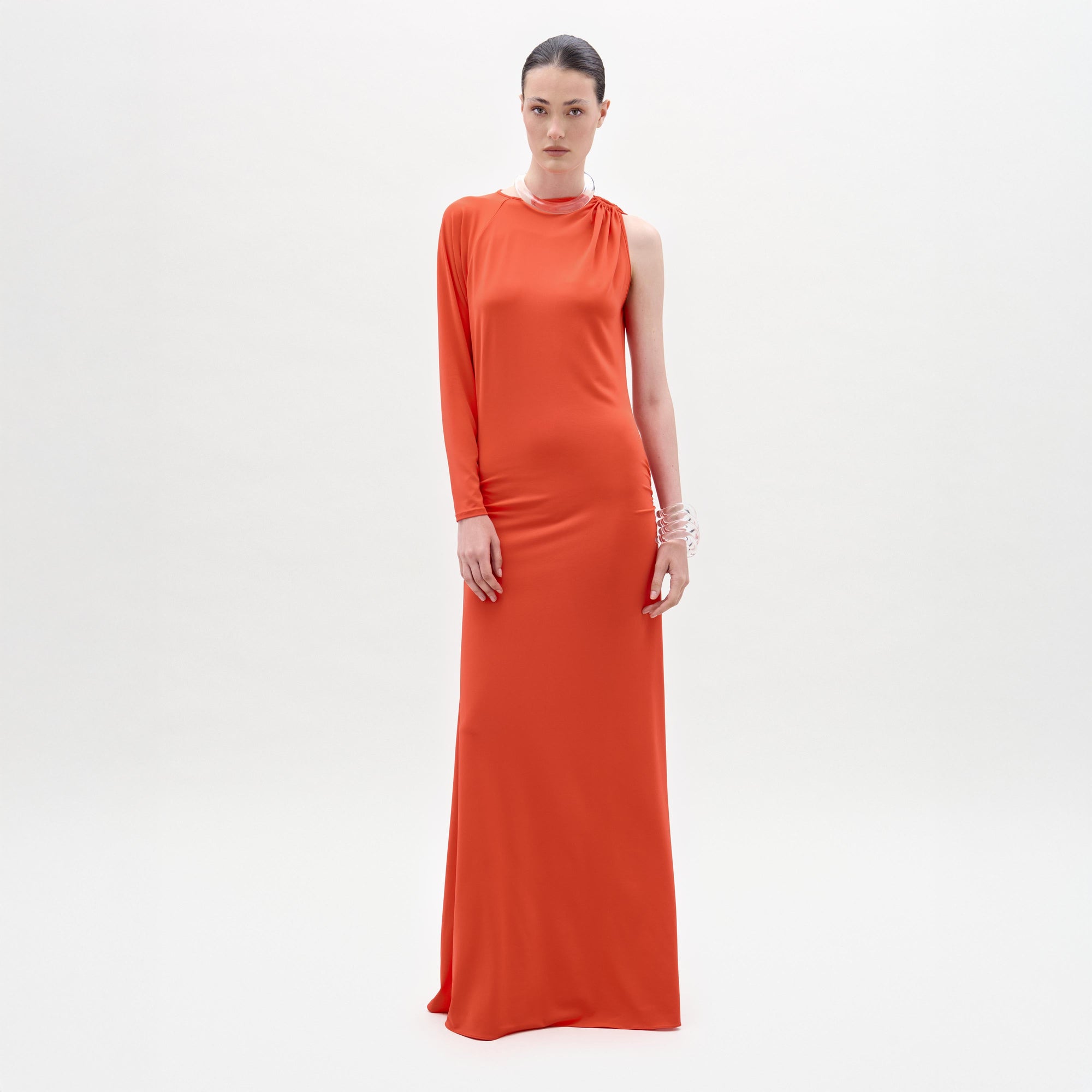 A woman stands against a plain white background, looking directly at the camera while wearing the Virna Dress—a long, one-sleeve orange maxi dress with a sleek column silhouette.