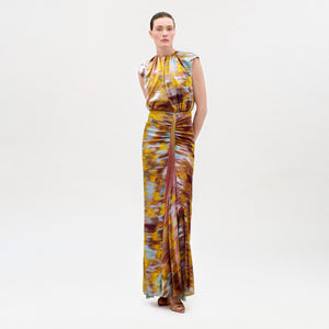 A woman stands against a plain background wearing the Winona Dress, a sleeveless, floor-length dress featuring a colorful tie-dye pattern in yellow, blue, and maroon tones, paired with brown sandals.