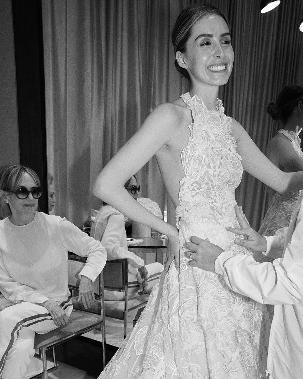 A woman smiles while wearing a detailed lace gown, with a person adjusting it. Another woman sits nearby watching.