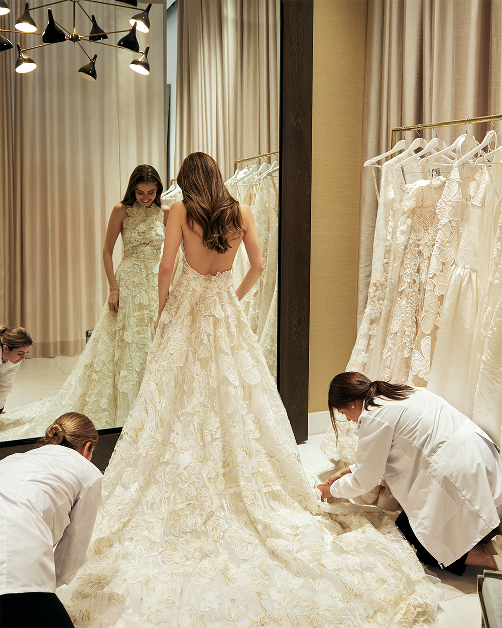A woman in a bridal gown stands in front of a mirror, with two assistants adjusting the train. Several other wedding dresses hang in the background.