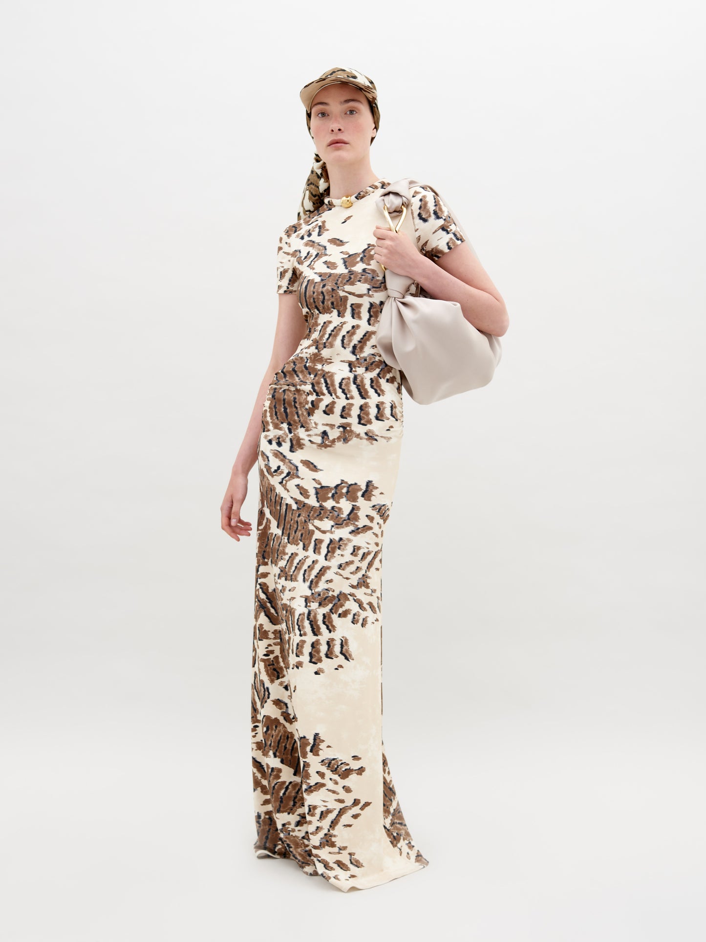 A person wearing a Yara Dress Beige Abstract Animal Print and cap with a white bag over their shoulder stands in front of a plain background, ready for pre-order by the November 15th, 2024 ship-by date.