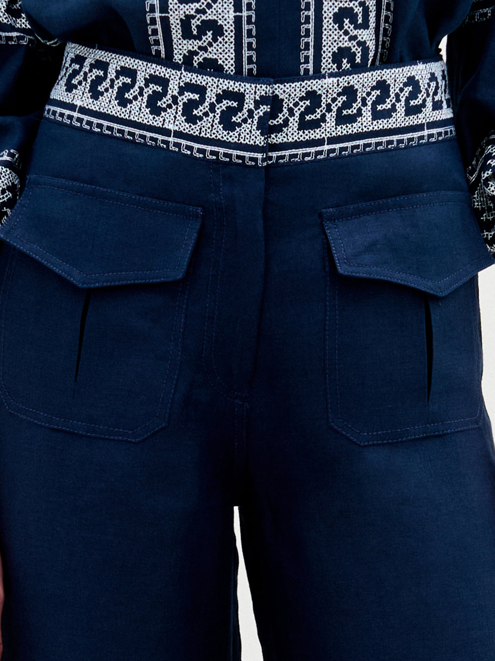 A woman stands against a plain background, wearing a navy blue embroidered blouse with ruffled sleeves and the Zina Pant—matching embroidered linen wide-leg pants—paired with gold sandals.