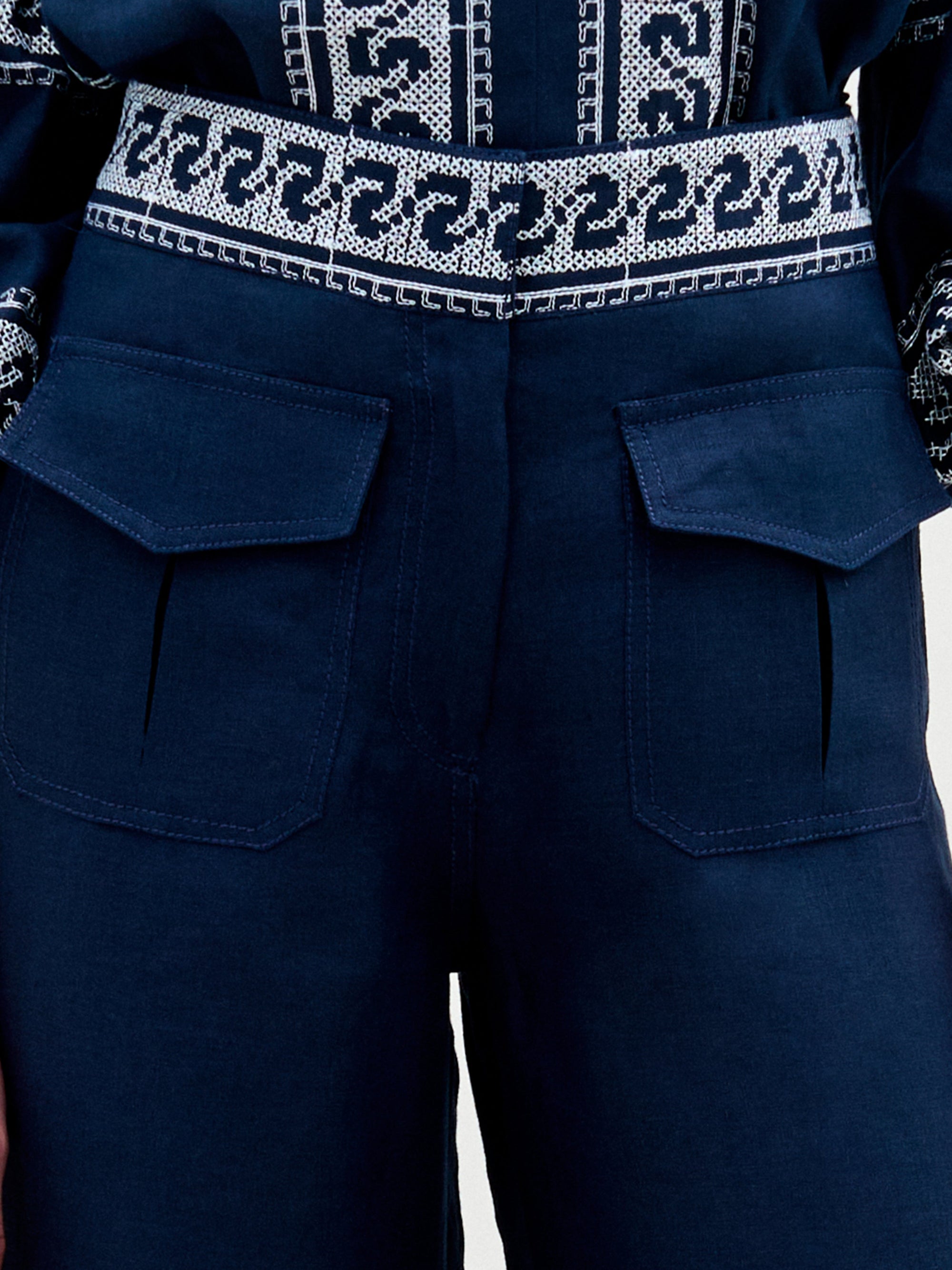 A woman stands against a plain background, wearing a navy blue embroidered blouse with ruffled sleeves and the Zina Pant—matching embroidered linen wide-leg pants—paired with gold sandals.