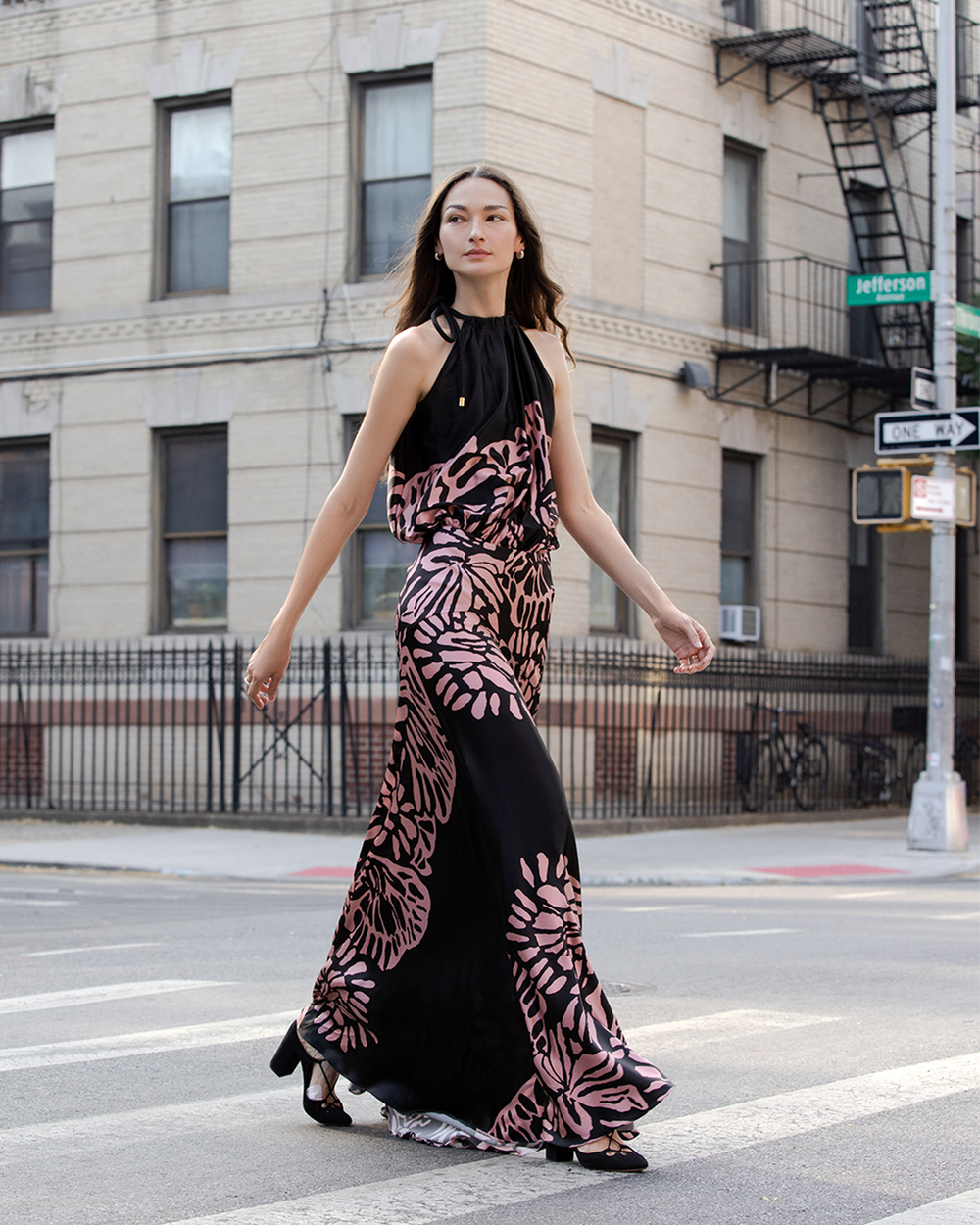 A person in a black and pink patterned dress walks confidently across a city street, with a beige brick building in the background.