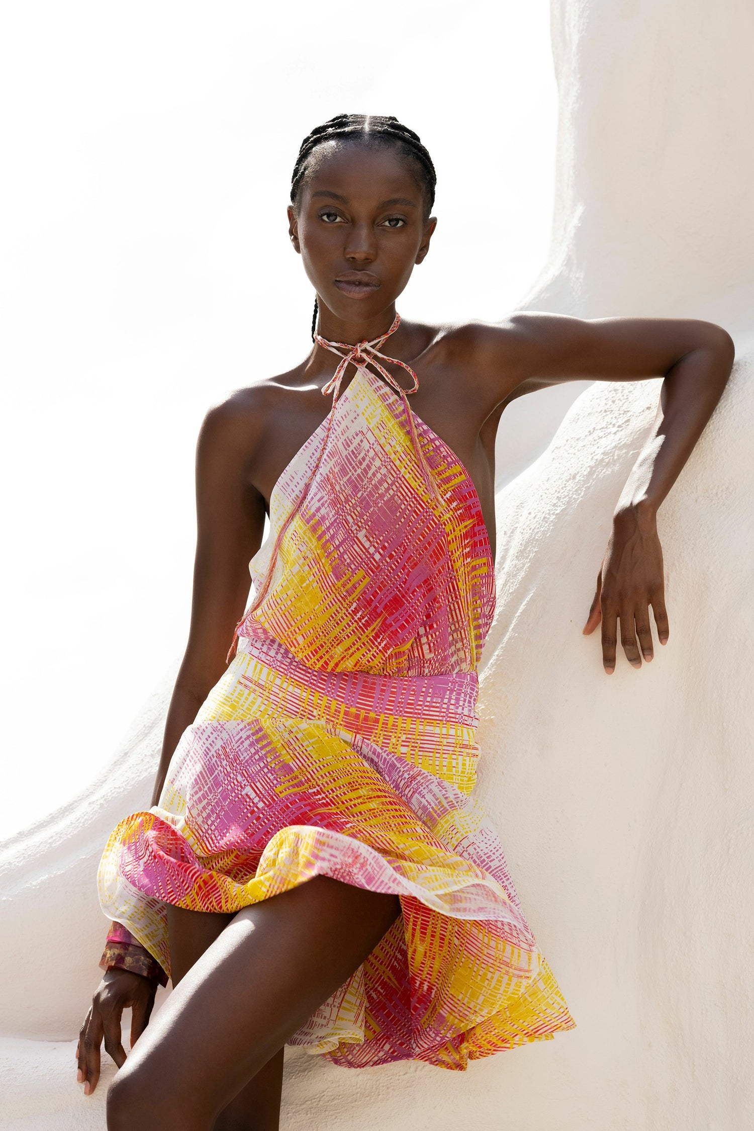 Person wearing a colorful, patterned halter dress leans against a white structure, with a bright sky in the background.