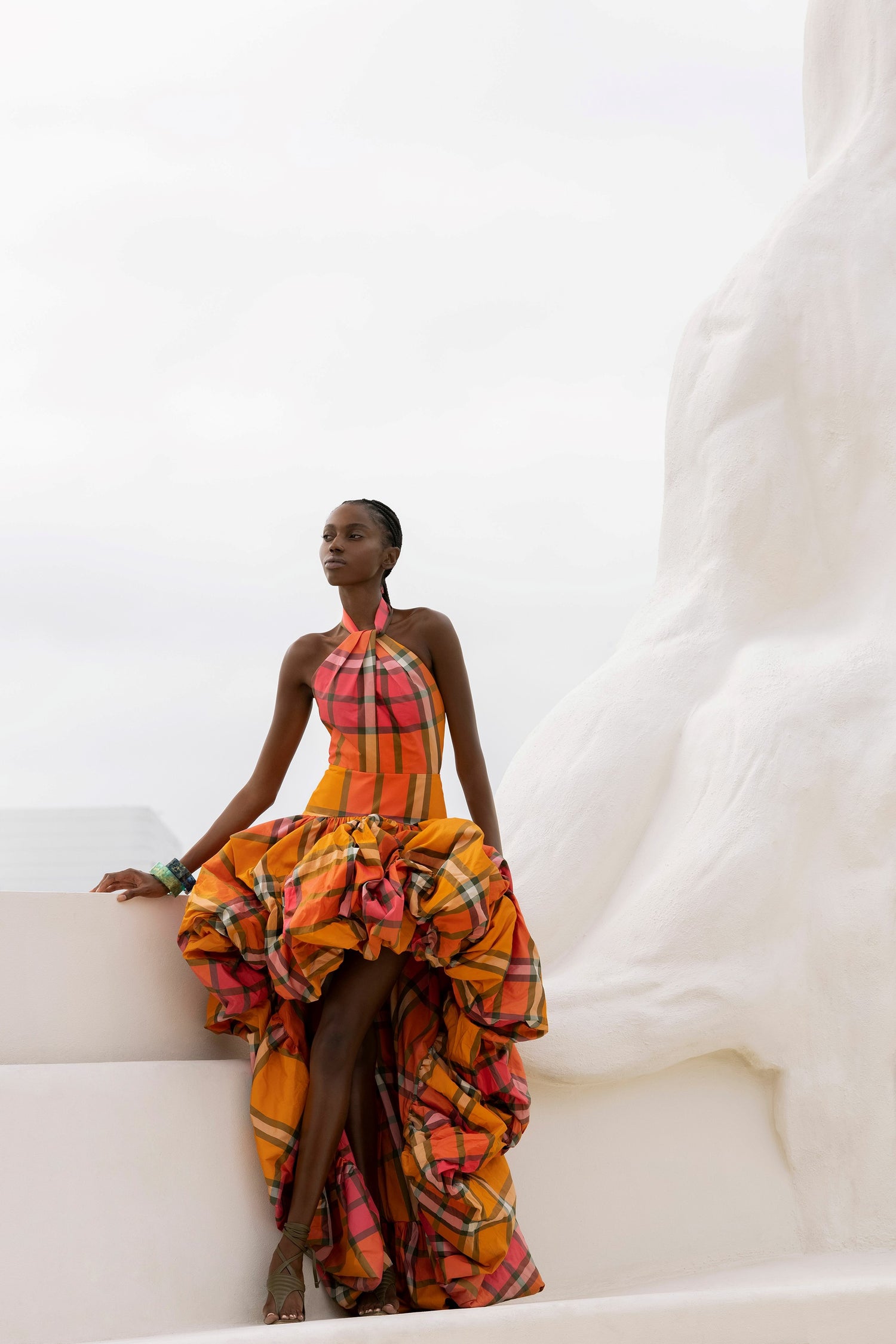 Person wearing a colorful, ruffled dress poses outdoors against a white structure under a cloudy sky.