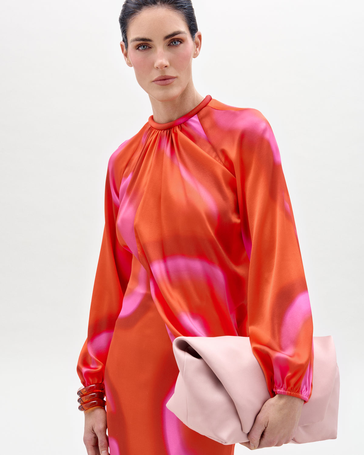 The supermodel Hillary Rhoda wearing a vibrant orange and pink dress holding a pink clutch against a white background