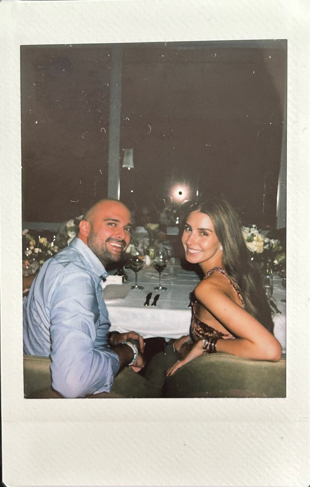 A man and woman seated at a dining table, smiling at the camera, with floral centerpieces and tableware visible.