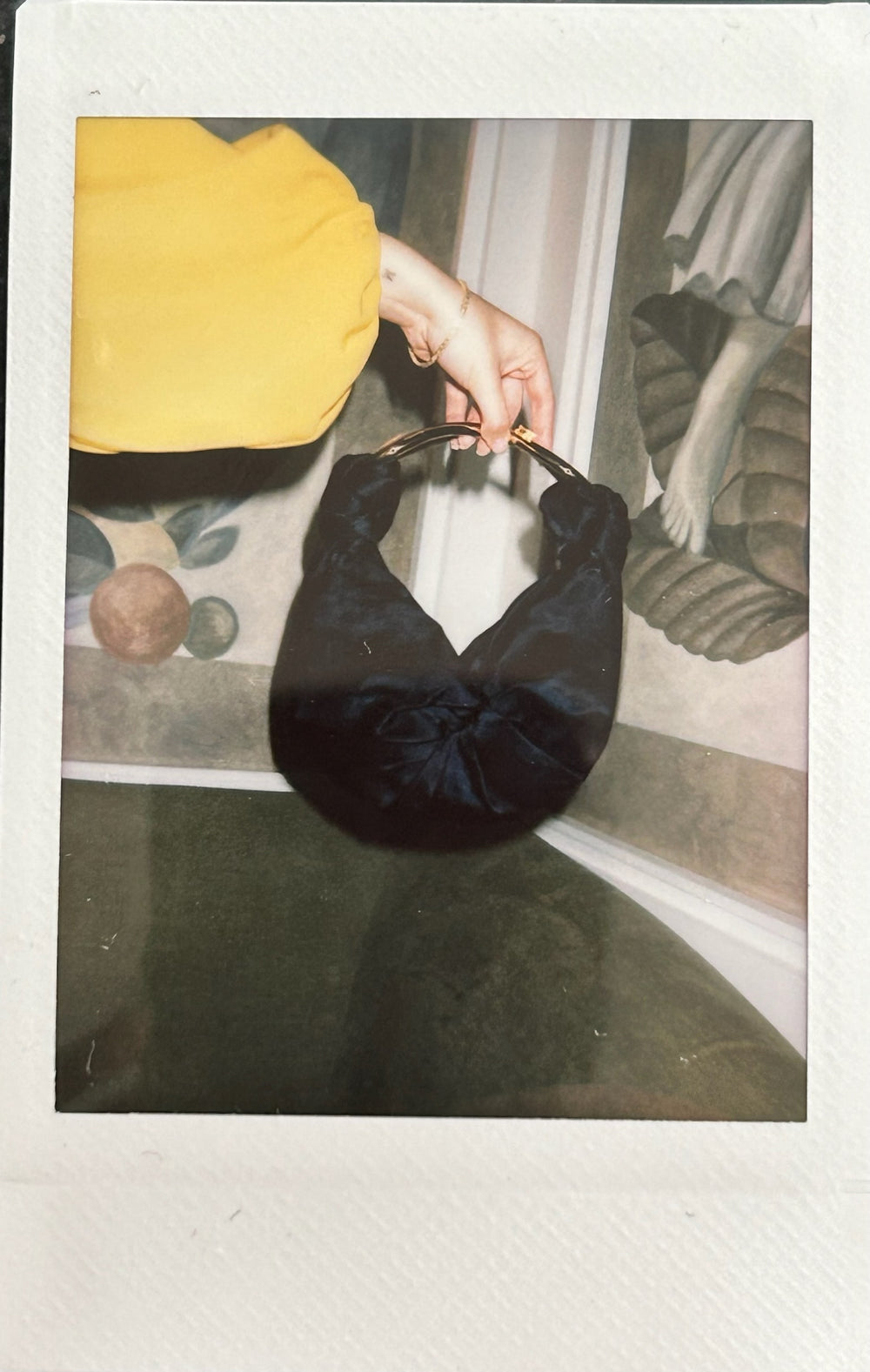 A hand in a yellow sleeve holds a black fabric bag with a circular handle in front of a wall with artistic designs.