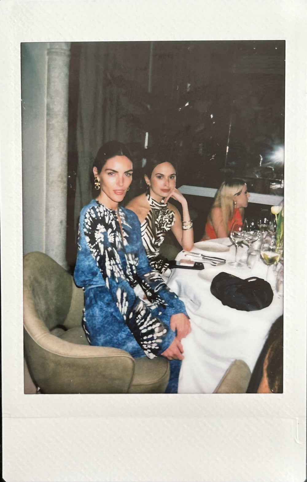 Two women sit at a dining table, dressed in stylish outfits. Empty glasses and a bottle of wine are on the table. Two other people are partially visible in the background.