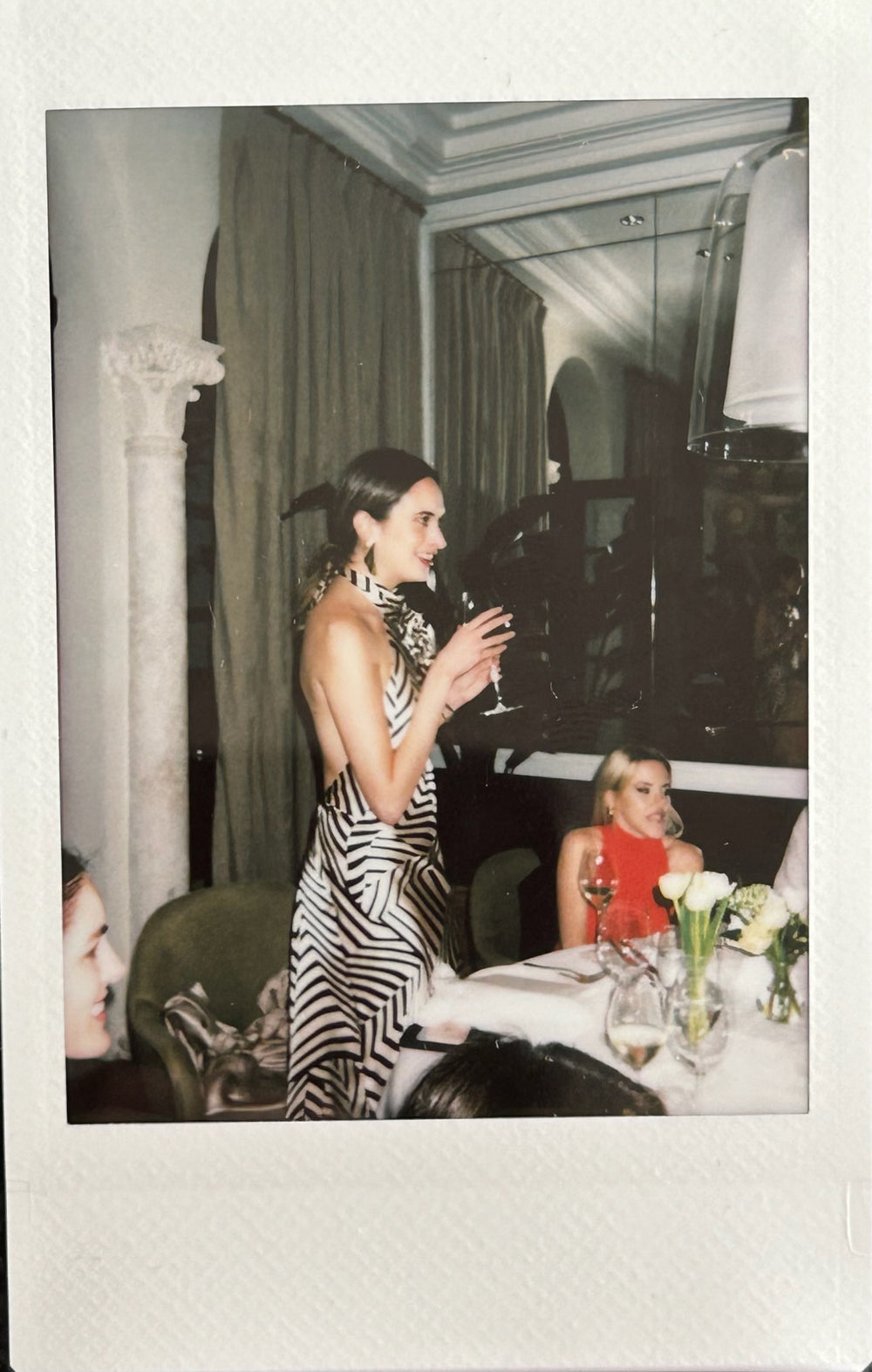 A woman in a zebra-patterned dress stands holding a wine glass at a dining table with seated guests in an elegant, draped room.