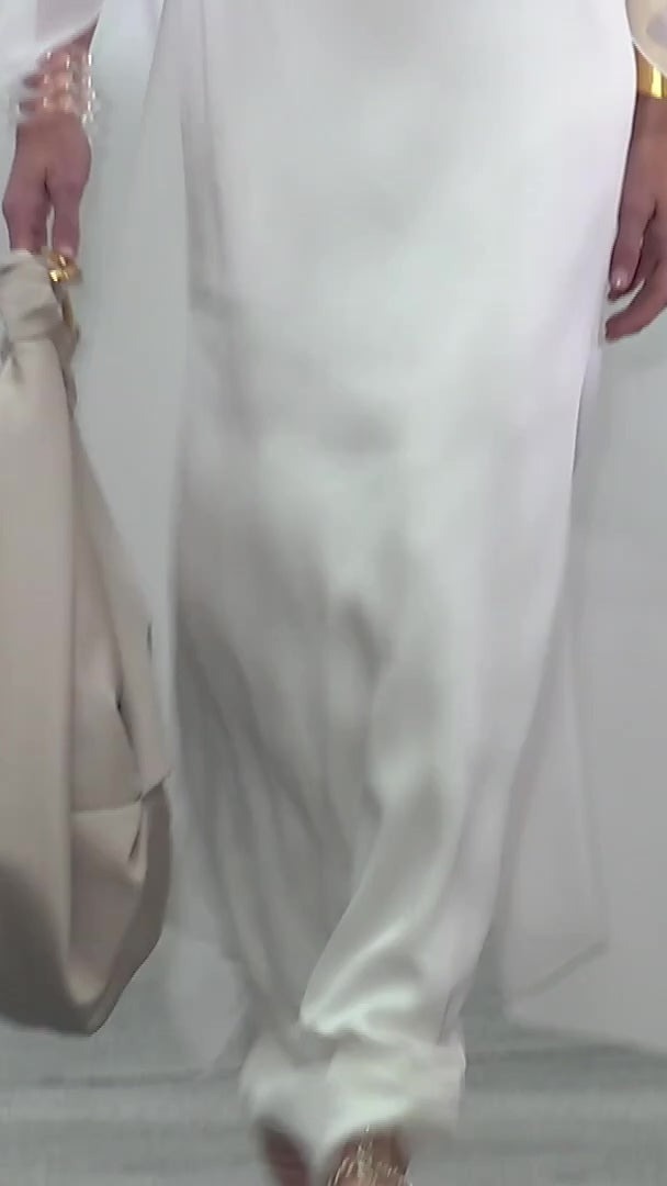 The supermodel Hillary Rhoda walking the runway wearing  the Kalina Dress, a floor-length, white sheer gown featuring a flowing cape sleeve and high neckline. Her hair is styled back as she gazes slightly to the side.
