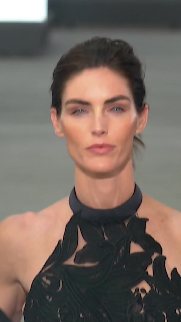The supermodel Hillary Rhoda walking the runway wearing the Mab Dress with large black floral appliqués over sheer nude fabric, featuring Italian textile cutouts at the waist and a high neckline.