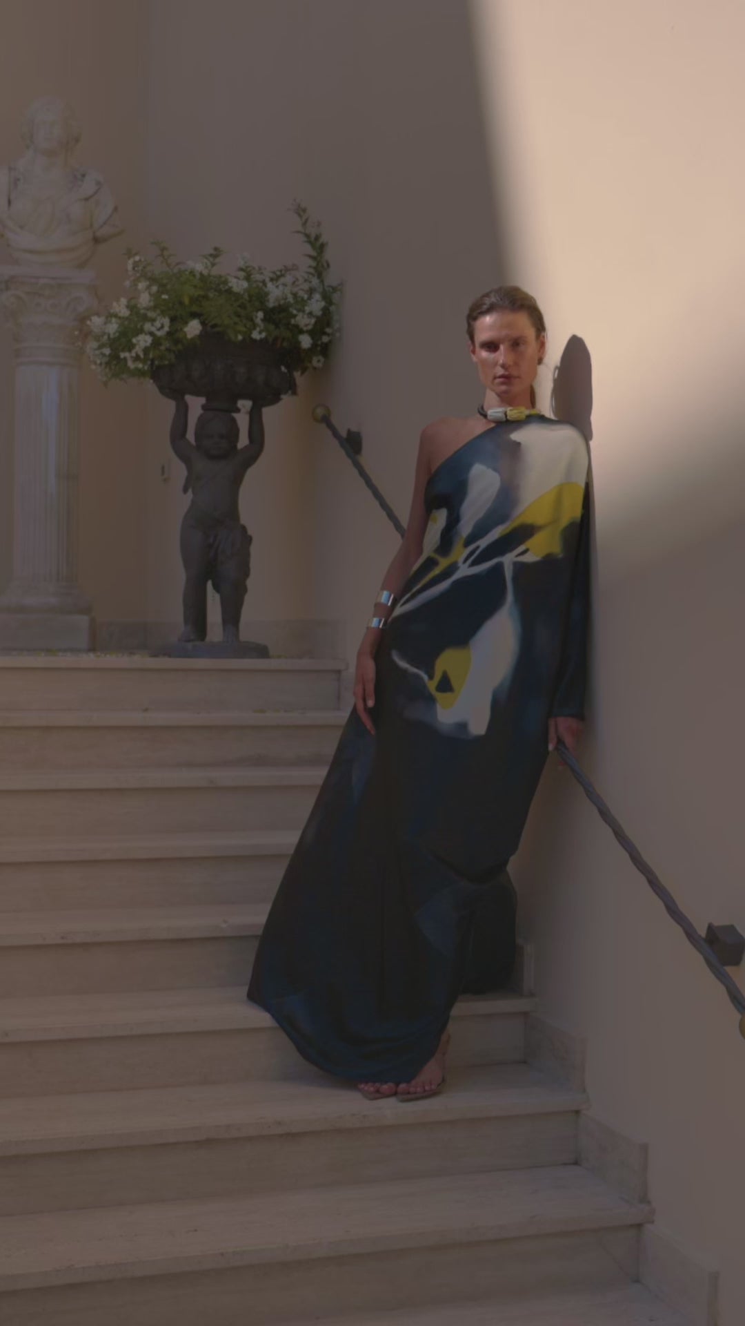 A women in a one-shouldered, abstract-patterned dress leans against a wall. They wear chunky jewelry and gaze thoughtfully, conveying a serene yet intense mood.