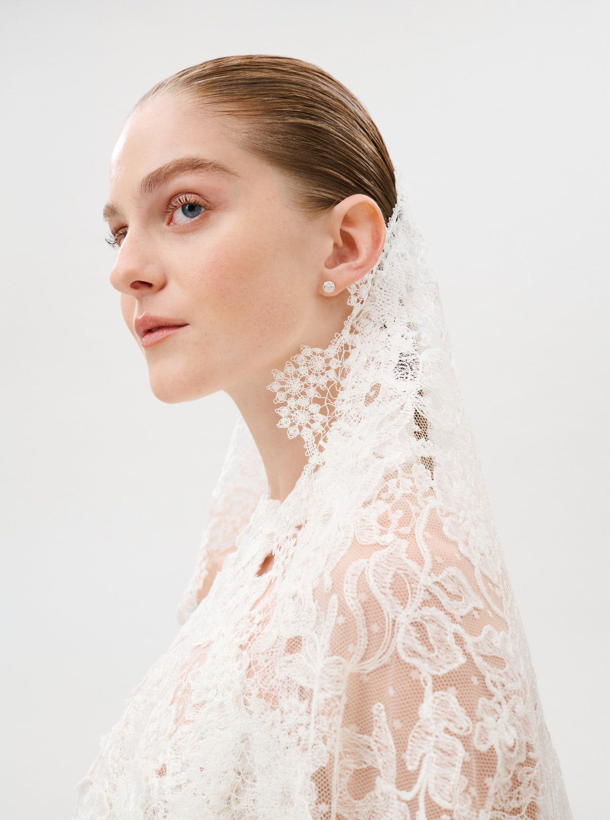 A woman wearing the Debra Veil, crafted from exquisite veil fabric, pairs it with delicate earrings as she gazes to the side. Her hair is slicked back, and she is dressed in a white lace garment against a plain background.