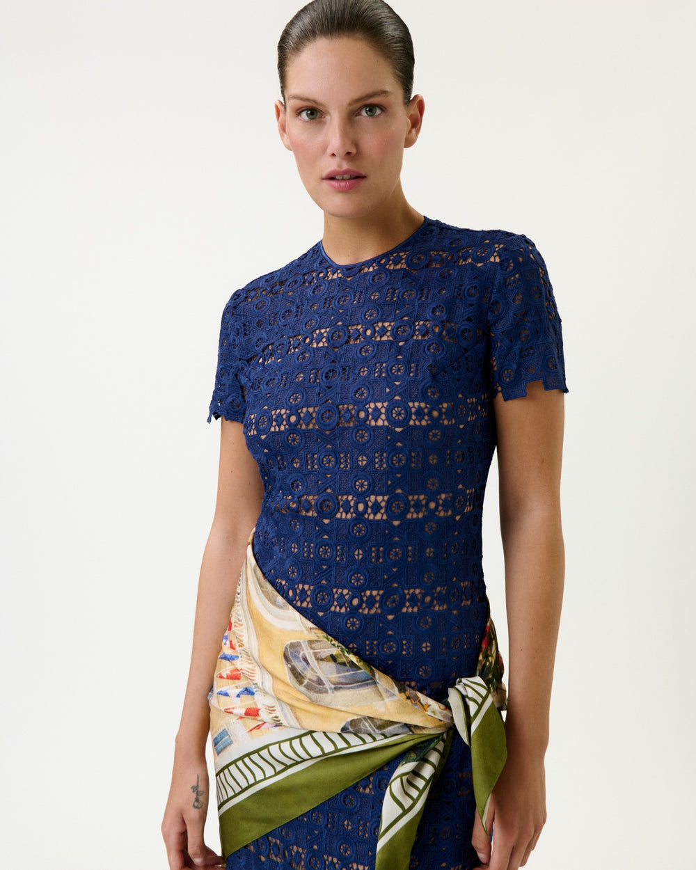 A woman stands against a white background, looking directly at the camera, wearing the Taiana Dress—a short-sleeve navy blue lace dress made from Italian guipure fabric—with a patterned scarf tied at her waist.