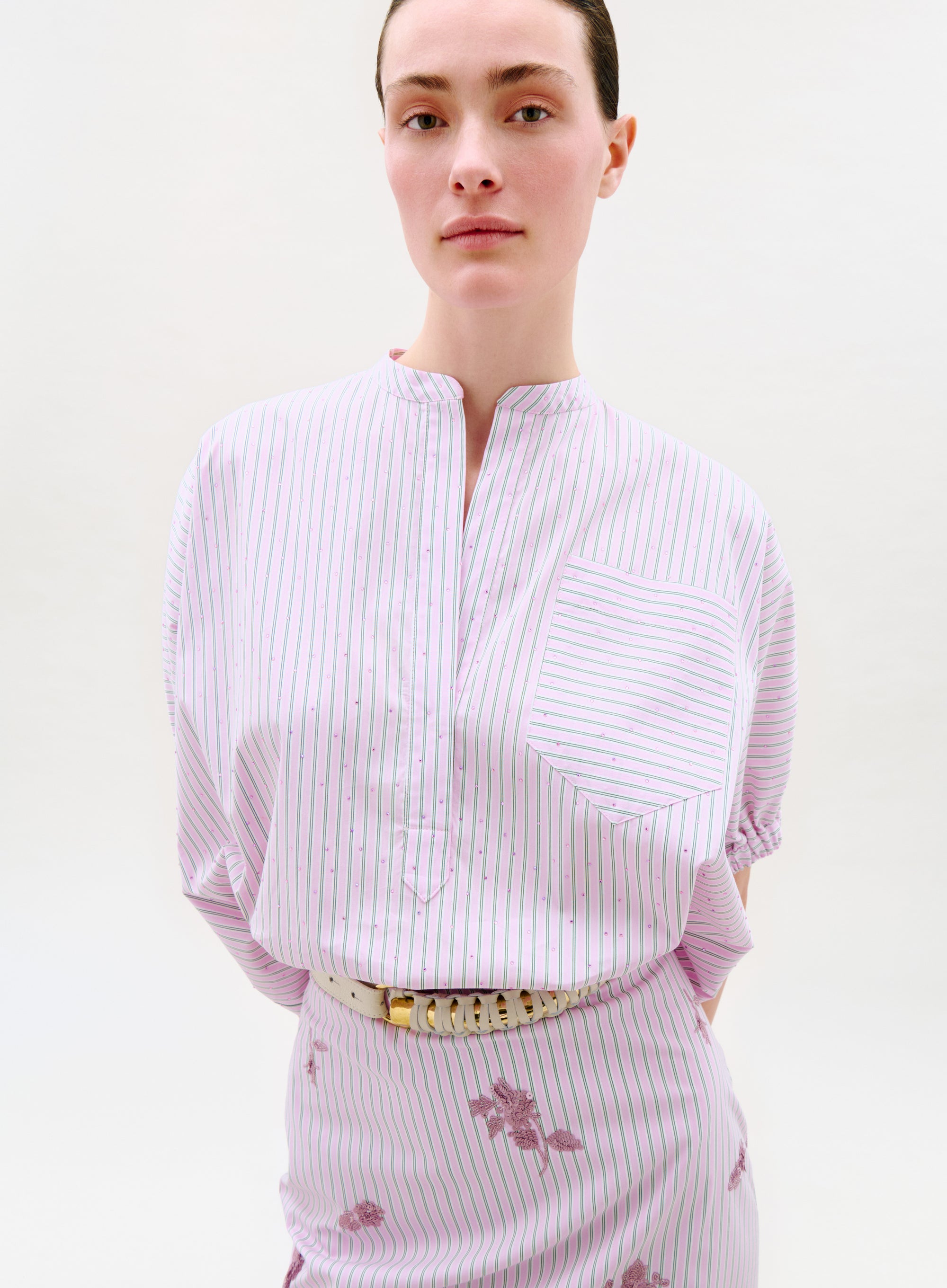 A person wears the Susane Blouse, a short-sleeve top made from Italian fabric with thin pink and green vertical stripes, a front pocket with horizontal stripes, a band collar, and a cream belt. The background is plain and light.
