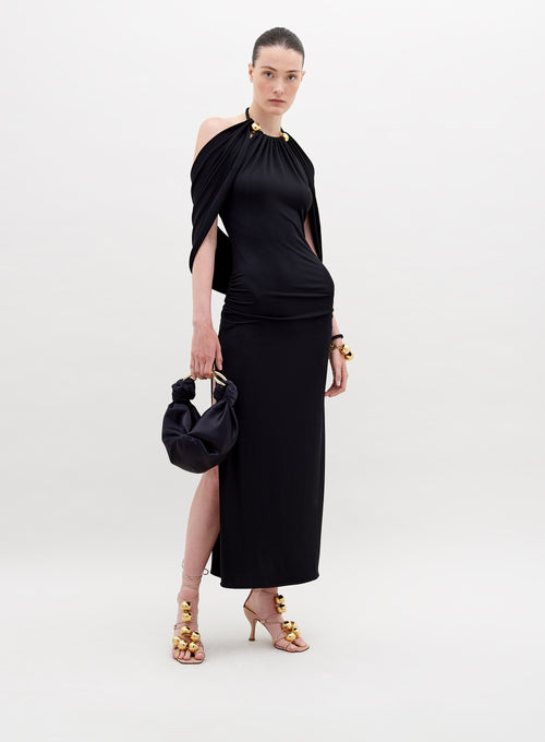 A woman stands against a white background, showcasing the Odele Dress Black with gold accents. She holds a small black bag and wears gold-embellished high heels. Pre-order now to ensure delivery by November 15th, 2024.