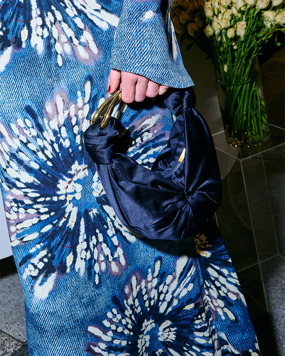Person holding a small, dark blue satin handbag with gold handles, wearing a blue patterned outfit.