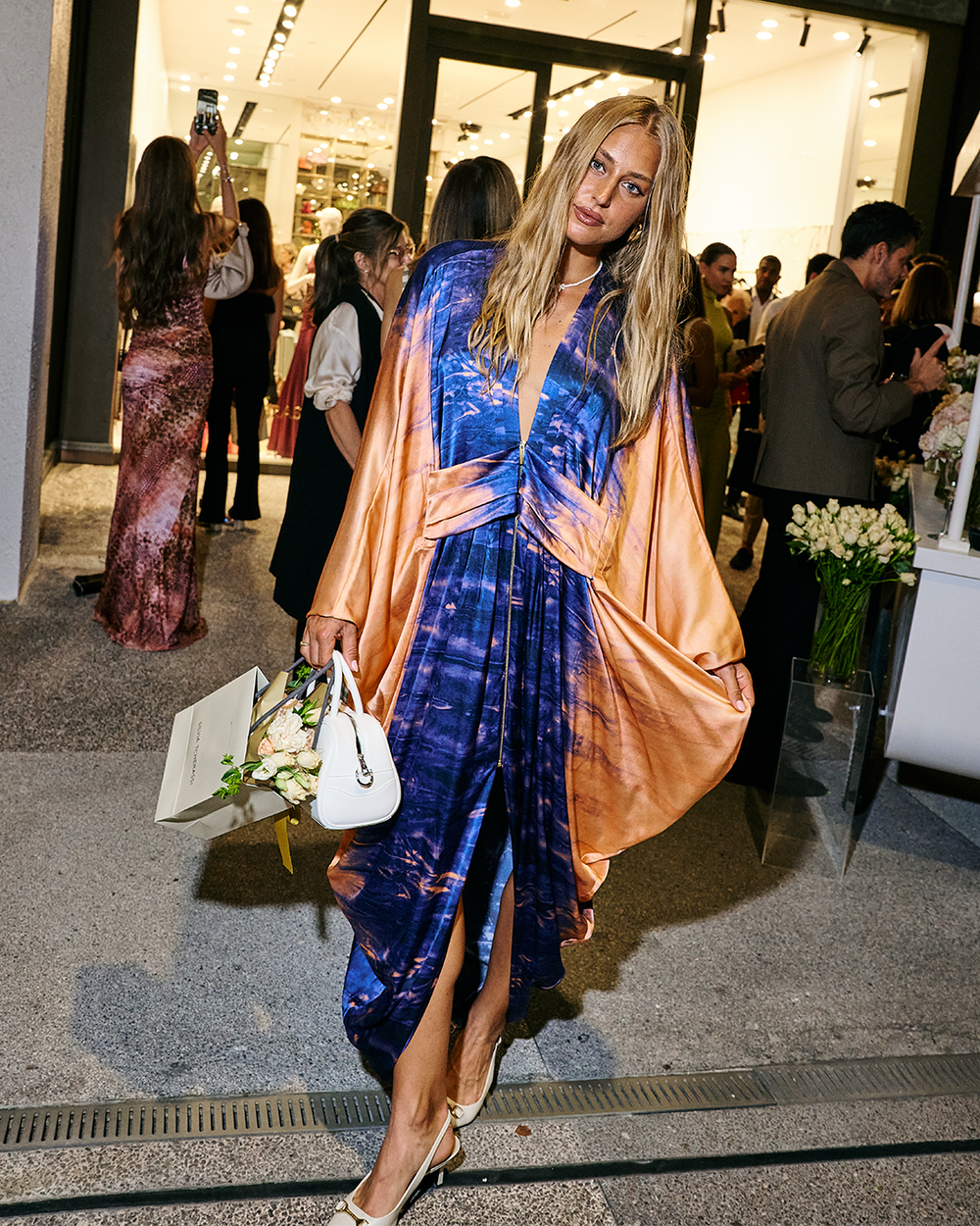 A person in a colorful dress poses outside a store holding a bouquet and a white bag, surrounded by a group of people.
