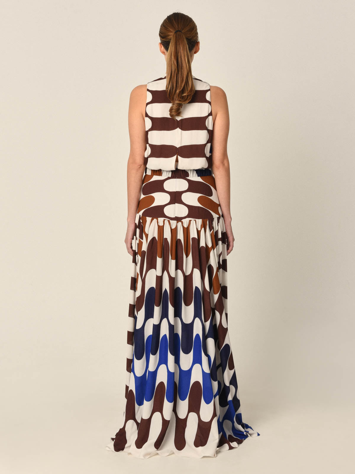 The Jael Dress Blue features long sleeves and a flowing skirt, adorned with an elegant geometric pattern in shades of brown, white, and blue that drapes gracefully to the floor.