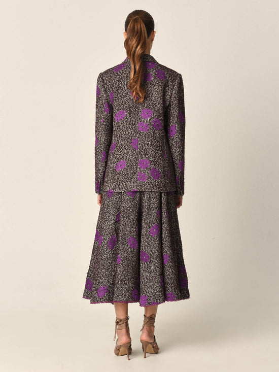 The Casilde Cape Violet is a brown textured cape adorned with violet gray floral embroidery, featuring a front tie closure with multi-color tassel details.