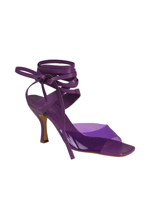 The Marlene Heels Purple, a high-heeled sandal with PVC transparent straps that tie around the ankle, are displayed against a white background.
