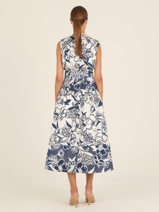 The Nayla Dress Navy Whimsical Garden is a sleeveless white dress with a deep V-neck and a flared skirt, boasting a blue floral print and a charming retro silhouette.